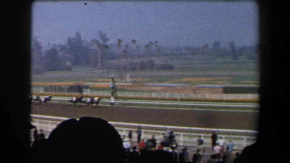 1958 California racetrack frame showing horses and jockeys running past the rail, with spectators and open grounds in view.