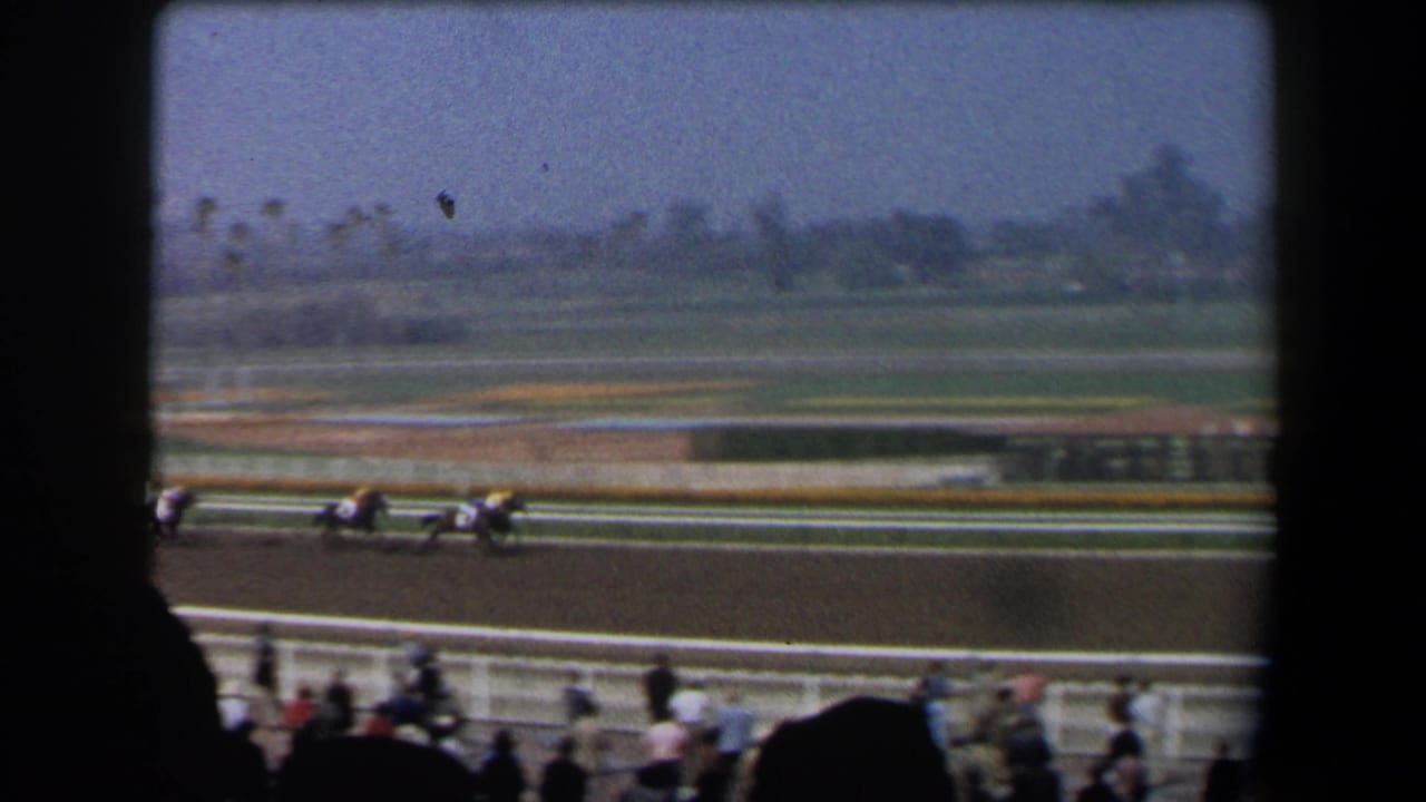 1958 California home-movie frame showing horses in mid-race on a dirt track, viewed from shaded stands with foreground spectator silhouettes.