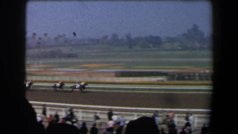 1958 California home-movie frame showing horses in mid-race on a dirt track, viewed from shaded stands with foreground spectator silhouettes.