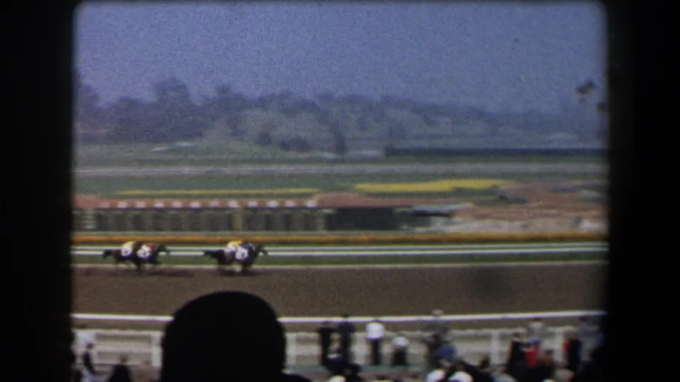 1958 California racetrack view with jockeys guiding horses at speed, seen across the rail from a spectator position.