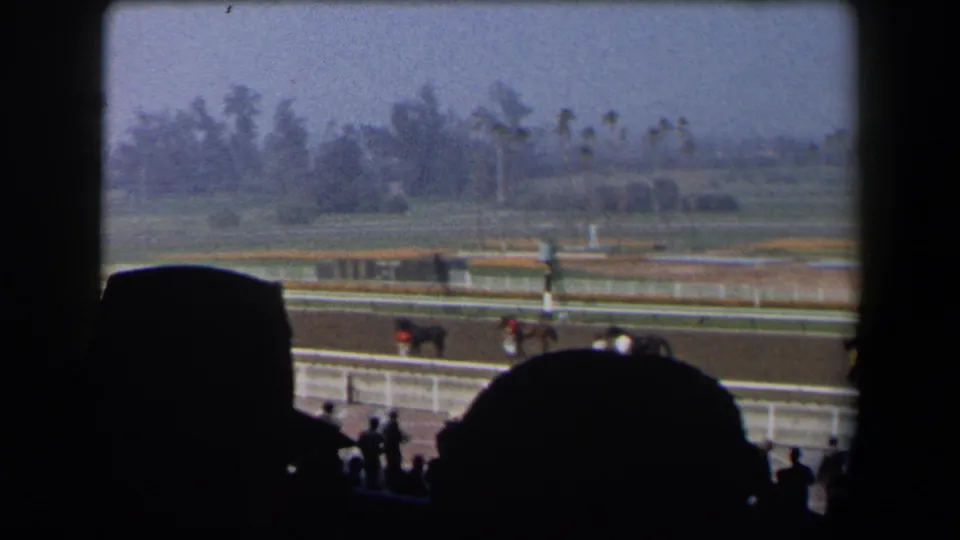 From the shaded stands, the 8mm frame looks across a California horse track with horses, riders, railings and landscaped grounds.