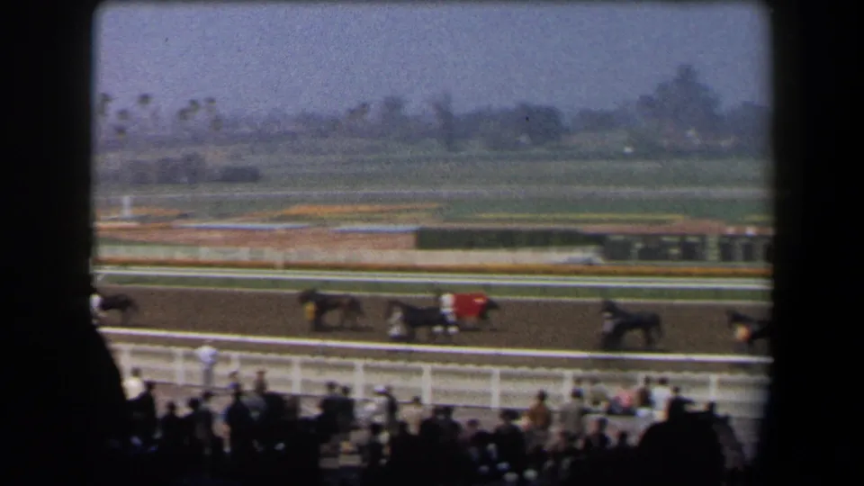 Late in the 1958 California racetrack clip, horses and riders move across the track below silhouetted spectators and a hazy infield.