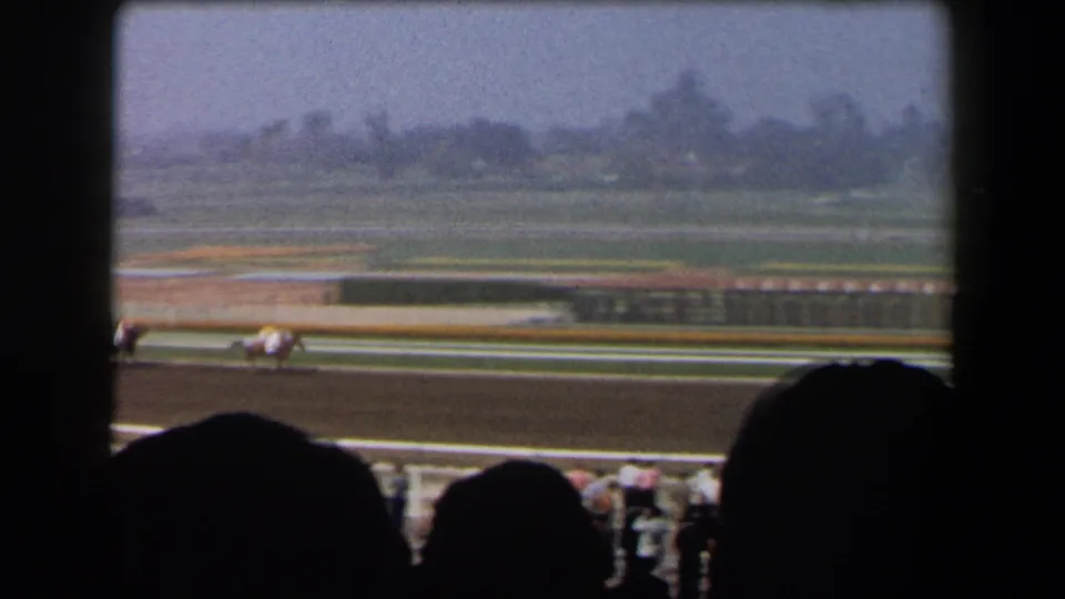 Horses and jockeys continue along a California dirt racetrack, seen past dark foreground spectators.