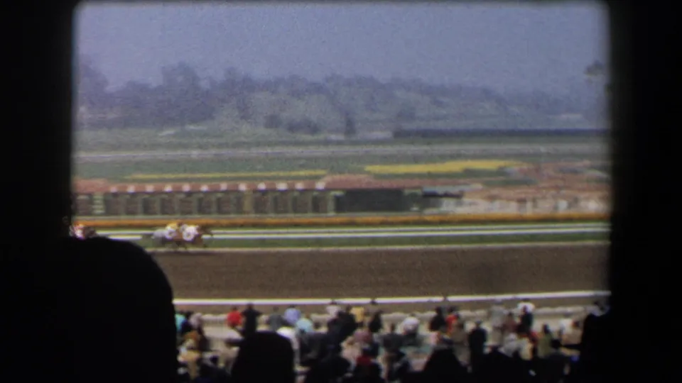 1958 home-movie frame of horses racing past spectators at a California track with hazy hills in the distance.