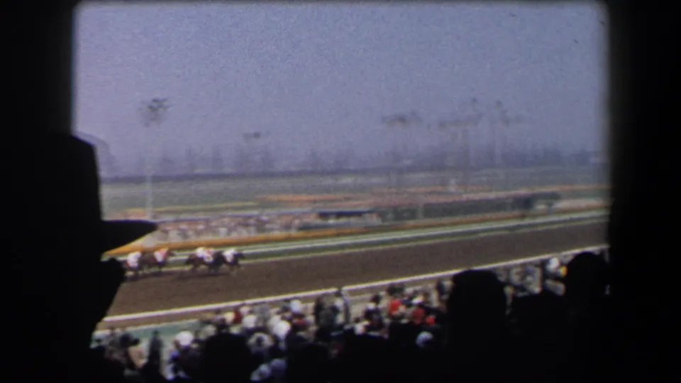 Horses and jockeys move along the racetrack as shaded spectators watch from a California grandstand in 1958 footage.