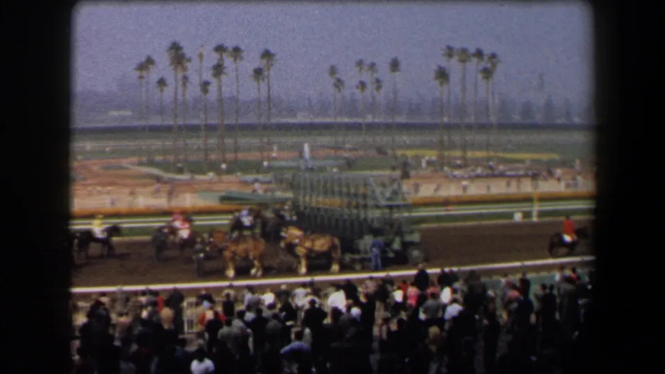 1958 California home-movie frame showing spectators in the stands as horses and jockeys wait near the racetrack starting gate.