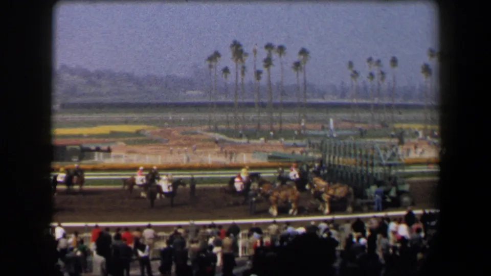 California 1958 home-movie frame of a horse race start area, with spectators in the stands and palm trees behind the track.