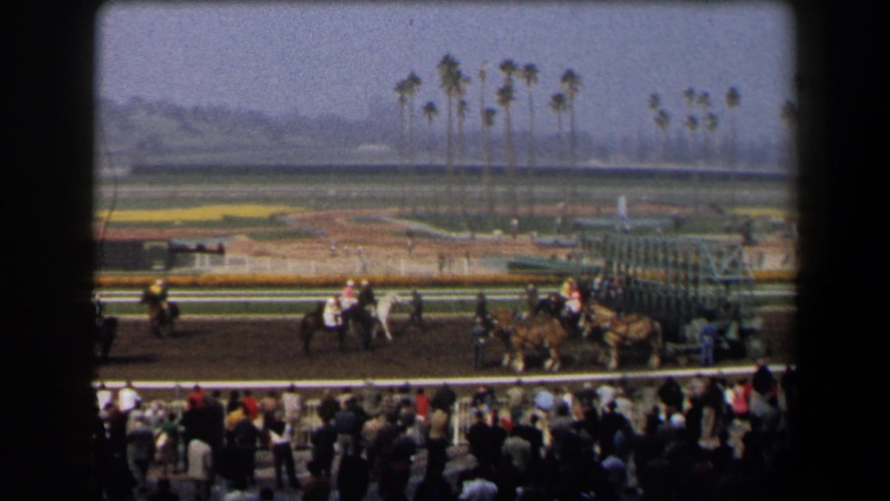 1958 California 8mm frame showing jockeys and horses on a racetrack near a starting gate, with spectators and palm trees visible.
