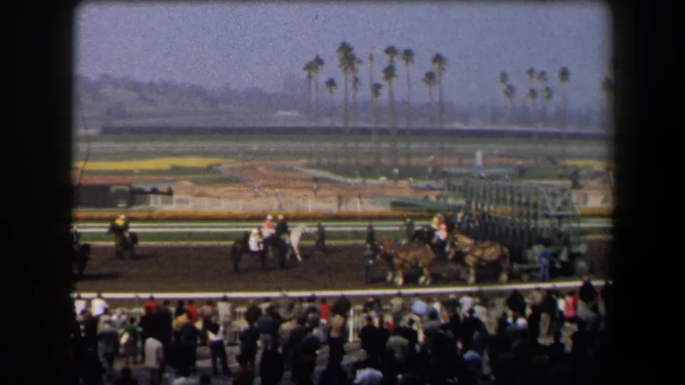 1958 California 8mm frame showing jockeys and horses on a racetrack near a starting gate, with spectators and palm trees visible.