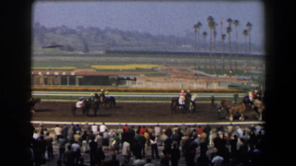 1958 California racetrack view with jockeys on horses passing the rail, spectators below, and palm trees across the infield.