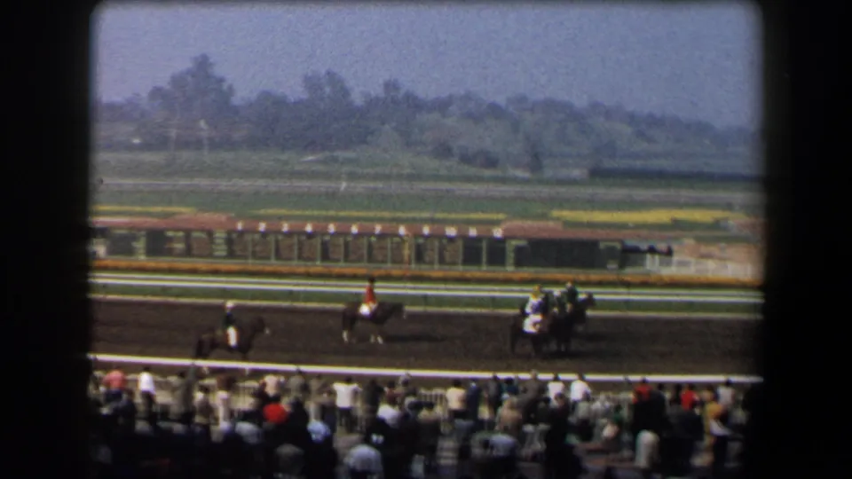 1958 California home-movie frame of a racetrack with jockeys on horseback, spectators, palm trees, and a distant landscaped infield.