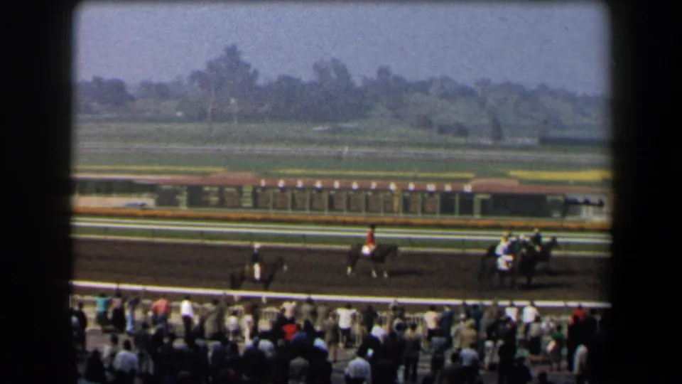 1958 California home-movie frame showing jockeys and horses on a dirt track as spectators watch from the rail.