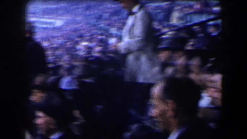 Soft, handheld 8mm crowd view from the grandstands, showing spectators seated and standing during the horse racing event.