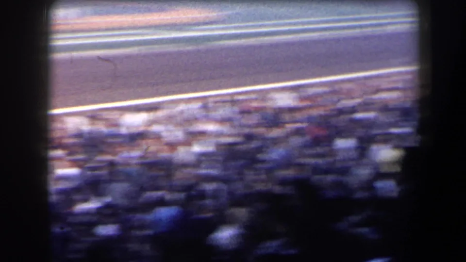 Handheld 8mm view across a crowded grandstand toward the racetrack, with motion blur and the track rail visible above the spectators.