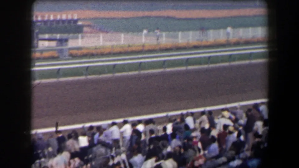1958 California 8mm frame showing a racetrack rail and seated spectators before the horses pass.