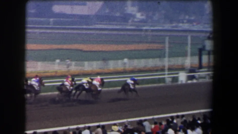 1958 California racetrack frame with several jockeys in colored silks riding past spectators.