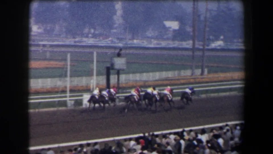 Home-movie frame of jockeys and horses bunched on a dirt racetrack, with crowd seating at the lower edge.