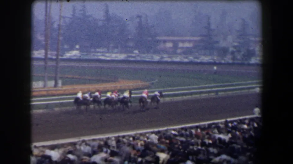 Wide 1958 California 8mm frame showing the racing field passing a packed spectator area under hazy light.