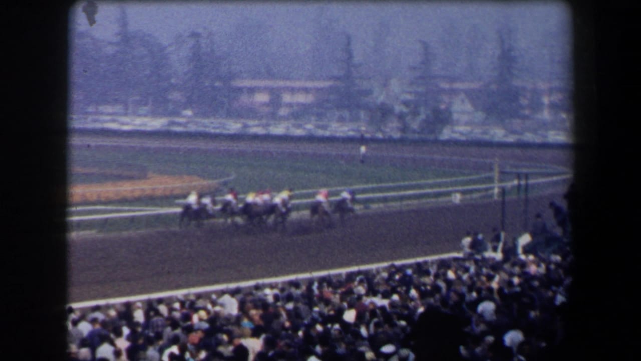1958 California 8mm view from the stands as racehorses and jockeys move along the dirt track before a dense spectator crowd.
