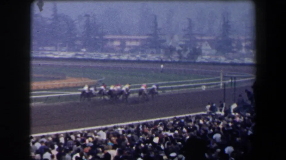 1958 California 8mm view from the stands as racehorses and jockeys move along the dirt track before a dense spectator crowd.