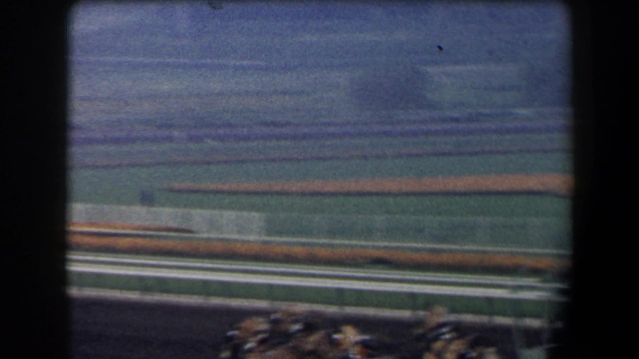 A racetrack curve and infield grass are seen with blurred horses entering the lower edge of the frame.
