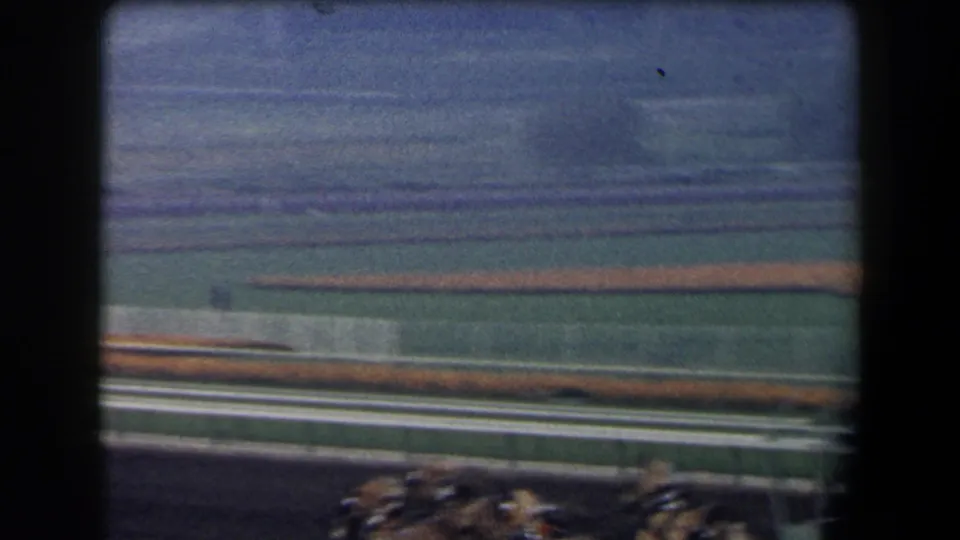 Wide 8mm racetrack view in California with the race briefly crossing the lower frame and open infield beyond the rails.