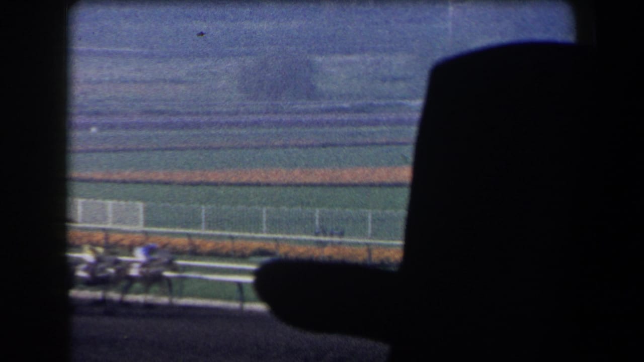 1958 California home-movie view of horses and jockeys racing past white railings, partly obscured by dark foreground structure.