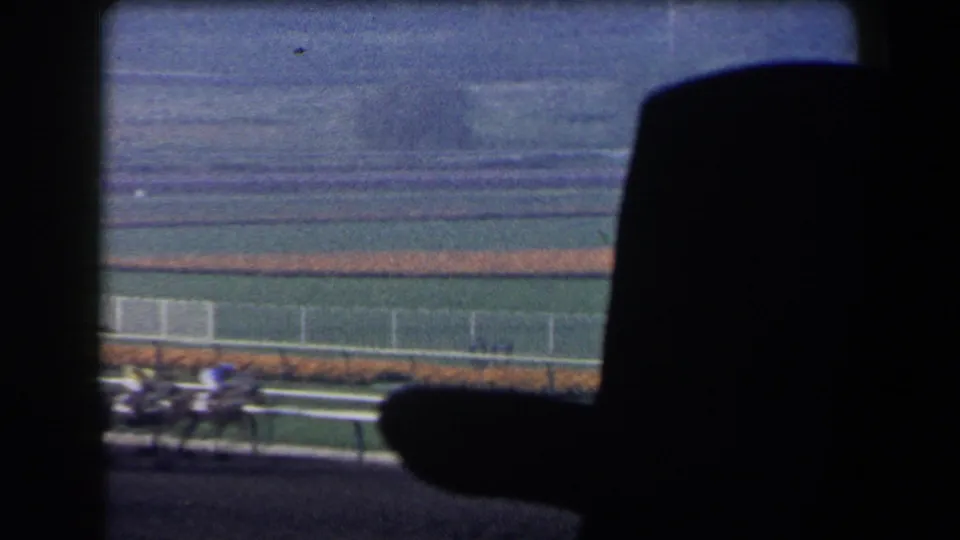 1958 California home-movie view of horses and jockeys racing past white railings, partly obscured by dark foreground structure.