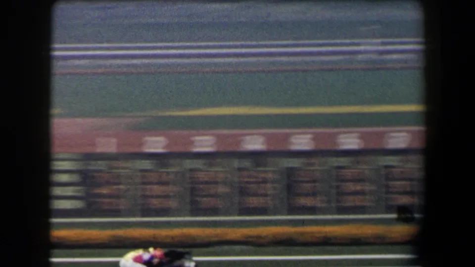 The camera sweeps across racetrack seating and railings, with racing horses partly visible near the bottom edge of the 8mm frame.