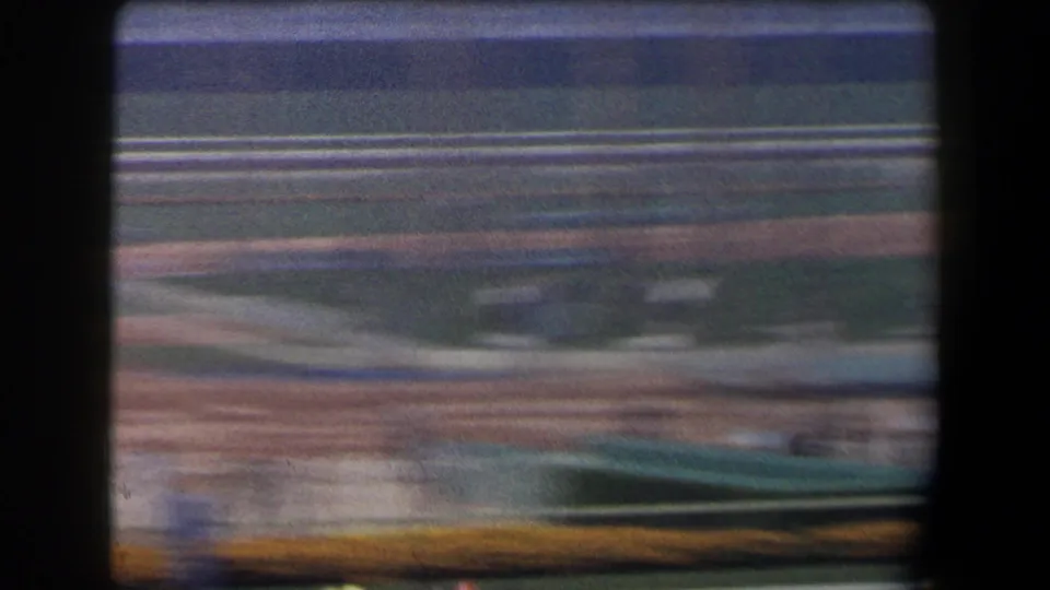 The 8mm camera catches a motion-blurred racetrack infield, with railings, turf, landscaping, and a dark obstruction at the frame edge.
