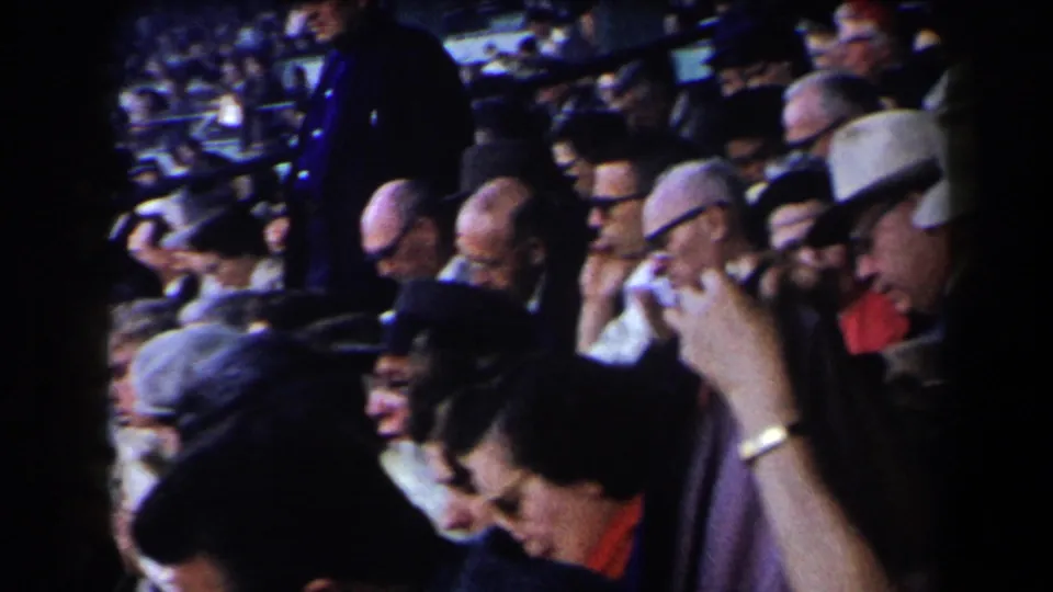 Adult spectators in hats, glasses, and coats sit shoulder to shoulder in a crowded 1950s California racetrack stand.