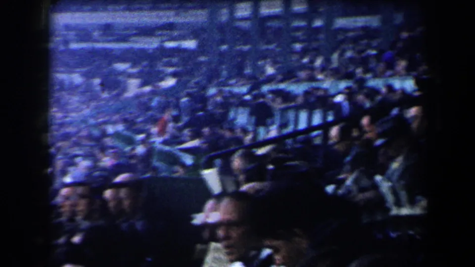 A handheld 8mm view looks across dense racetrack seating, with adults in hats and coats gathered behind railings.