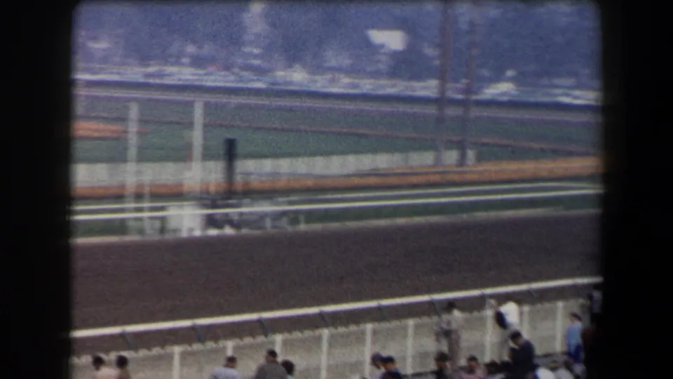 Home-movie frame of racetrack railings and track surface, with blurred motion and spectators along the lower edge.
