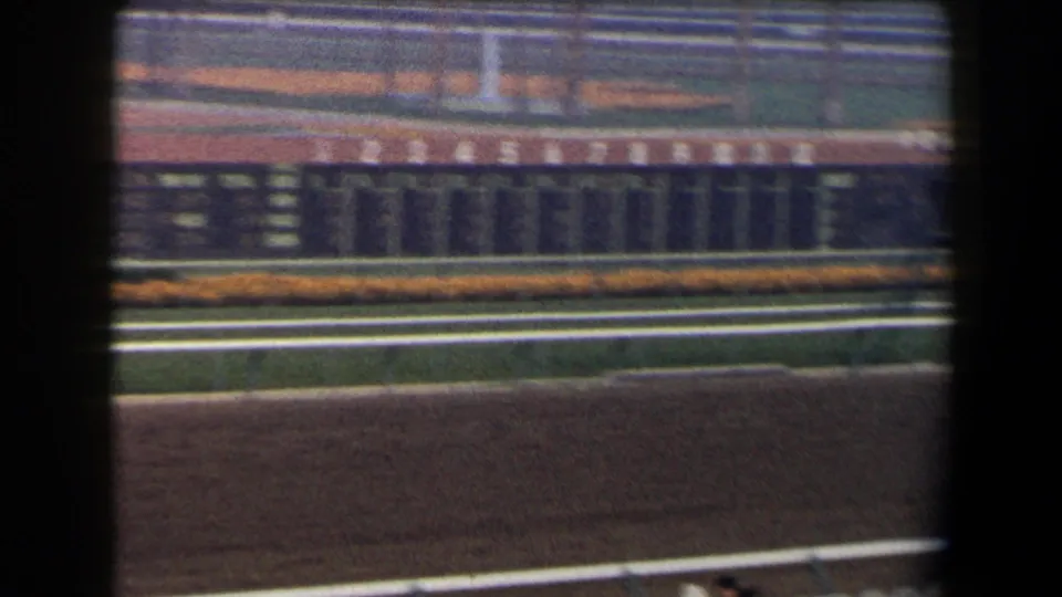 1958 California 8mm view across a horse racing track, with railings, infield, distant grandstand seating, and dark foreground obstructions.