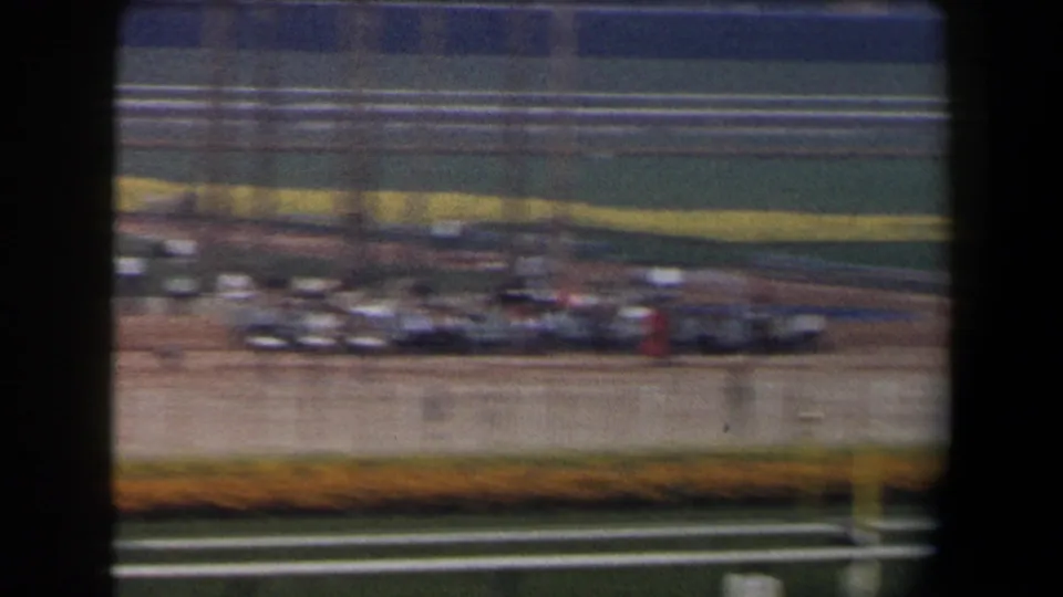 Motion-blurred 1958 California 8mm pan across the racetrack, showing white rails, yellow infield planting, and small distant figures.
