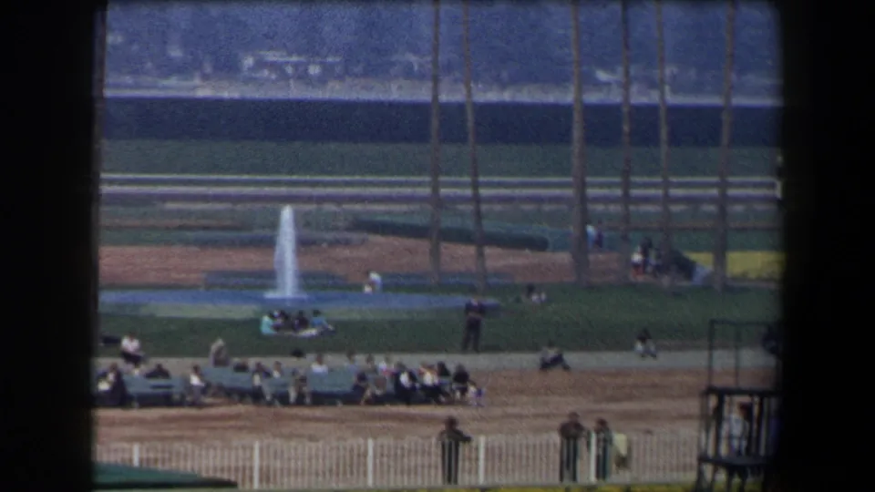 1958 California home-movie view of spectators beside a sunlit racetrack infield with a fountain, white railings, and palm trees.