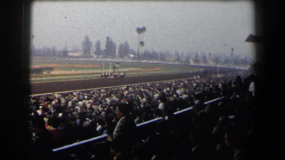 Packed California racetrack stands frame a distant group of horses running under a hazy sky in 1958 footage.