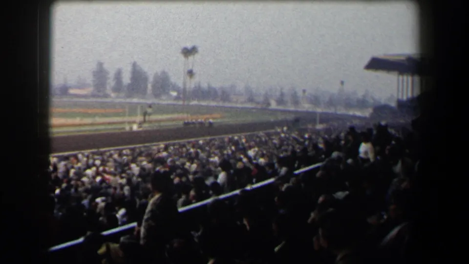 Home-movie angle from the stands shows a large crowd, track railings, and horses racing across a California course.