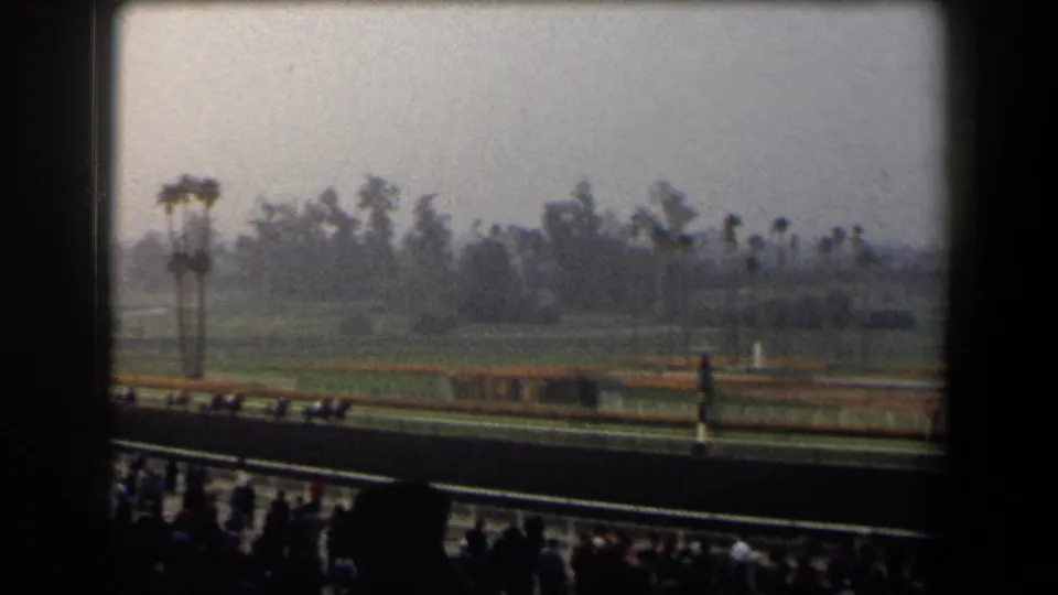1958 California racetrack view from the stands, with horses and jockeys moving along the track under a hazy sky.