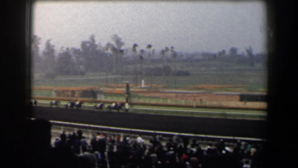Racehorses and jockeys move across a California racetrack while spectators watch from the lower foreground.
