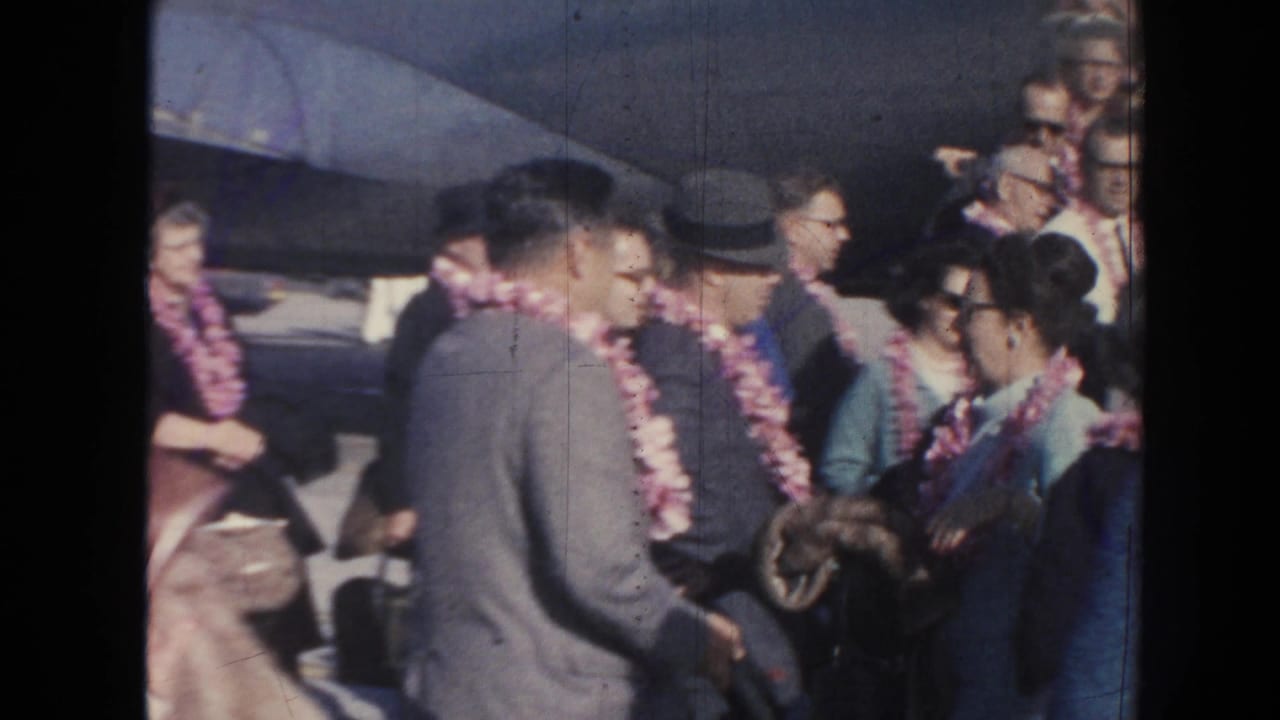 In the supplied 1961 Hawaii home-movie context, adults in suits and dresses wear leis and greet one another beside an aircraft at an airport arrival area.