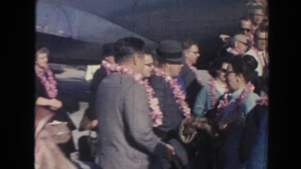 In the supplied 1961 Hawaii home-movie context, adults in suits and dresses wear leis and greet one another beside an aircraft at an airport arrival area.