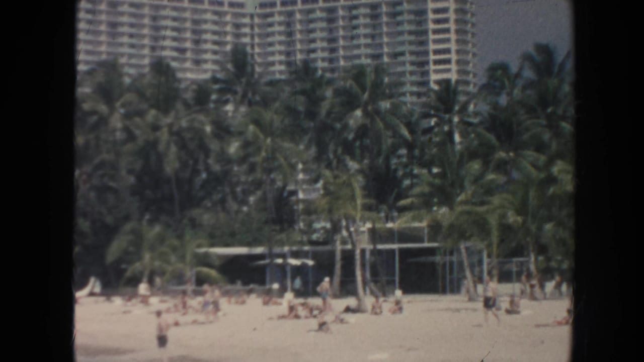 Hawaii beach view with a large resort hotel rising behind dense palms while people gather on the sand in the foreground.