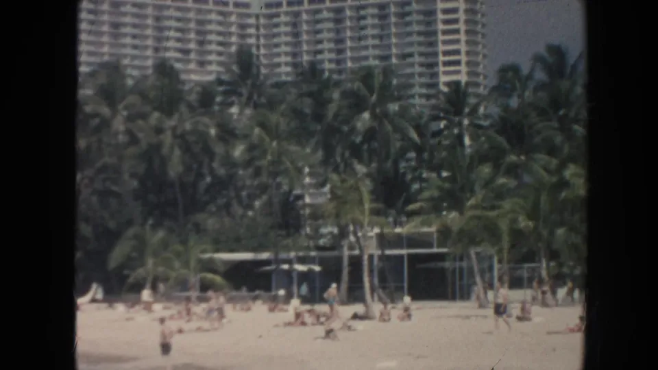 Hawaii beach view with a large resort hotel rising behind dense palms while people gather on the sand in the foreground.