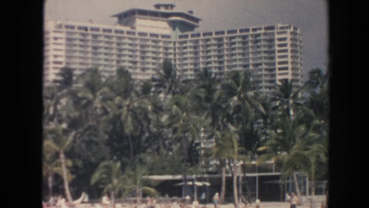 1961 Hawaii resort shoreline with a multi-story hotel, palm trees, beach structures, and distant people on the sand.
