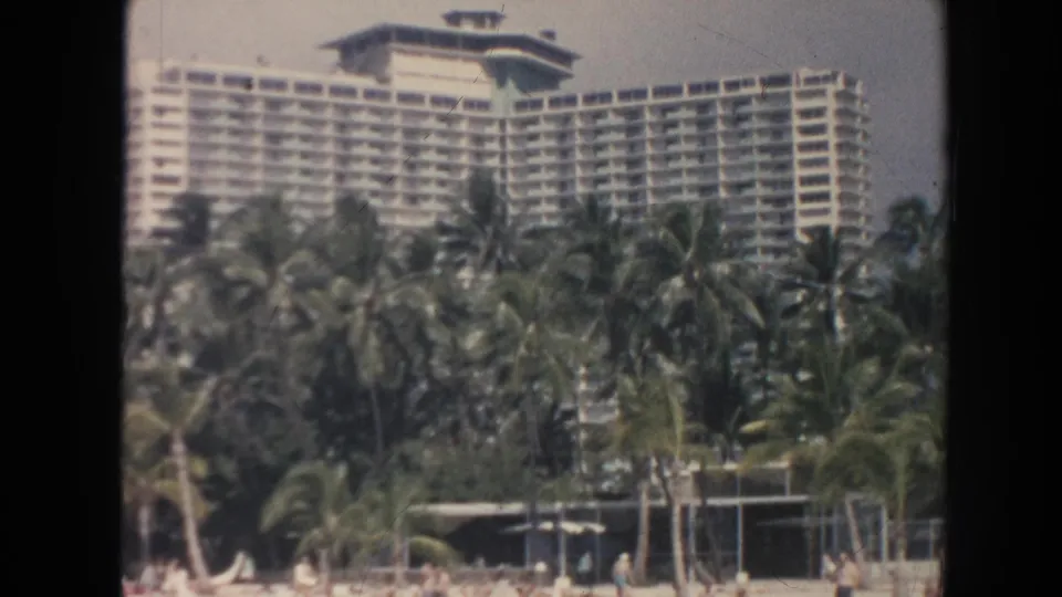 1961 Hawaii resort shoreline with a multi-story hotel, palm trees, beach structures, and distant people on the sand.