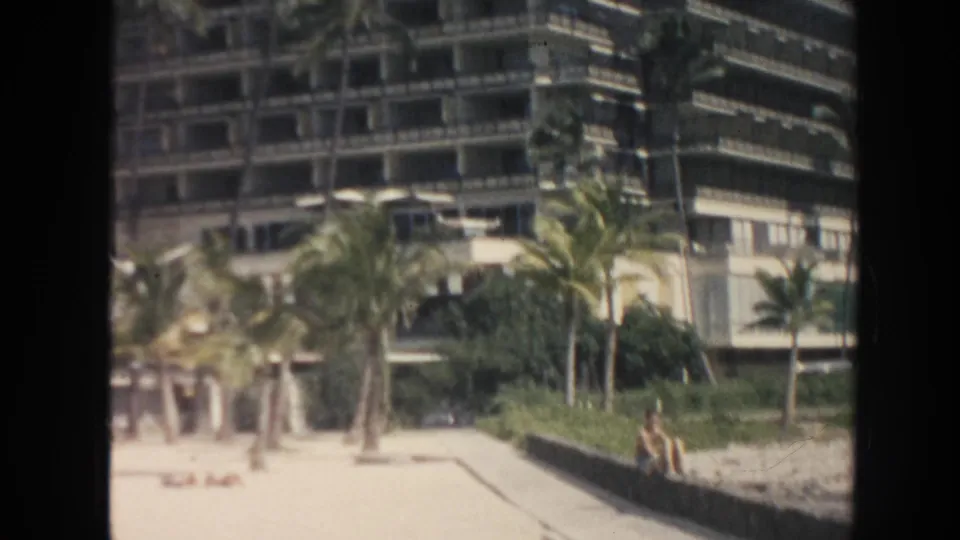 1961 Hawaii beach view with a tall resort building, palms, landscaping wall, and distant beachgoers.