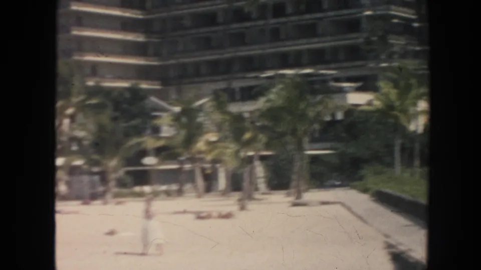 1961 Hawaii 8mm frame showing beach activity below a multi-story resort building and palm trees.