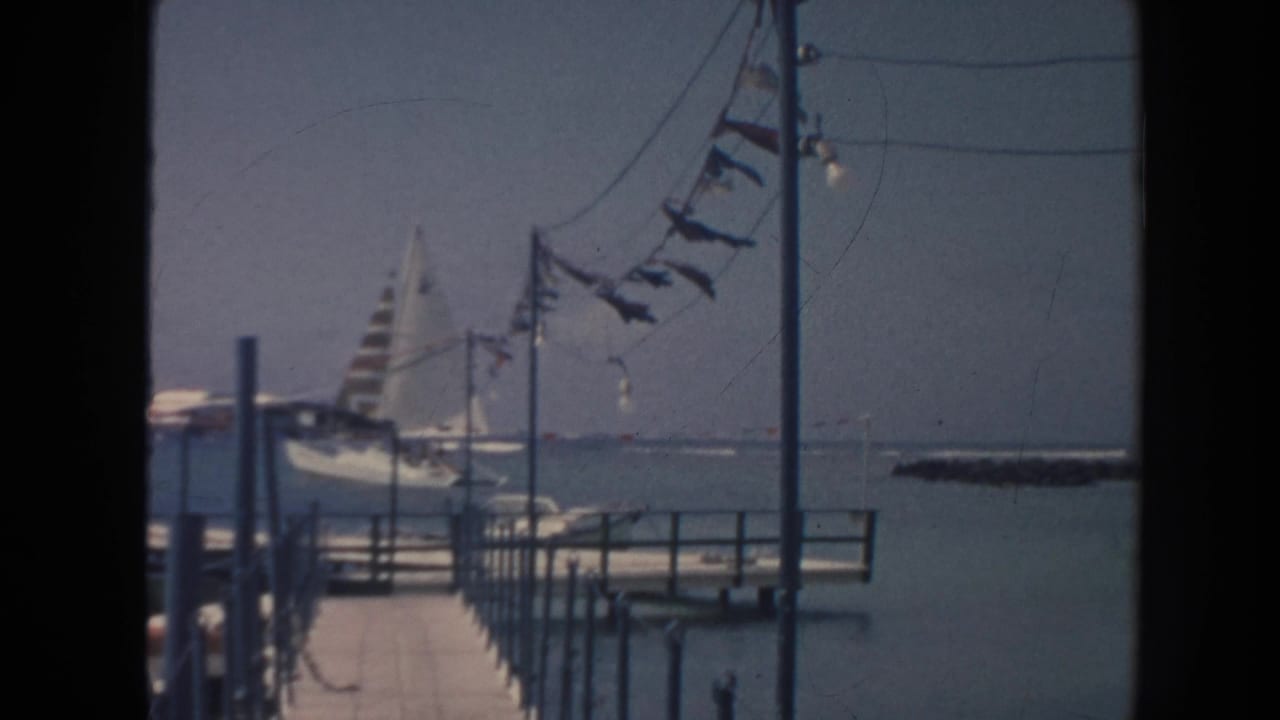 Pier beside calm water with a striped sailboat and pennant flags in the distance.