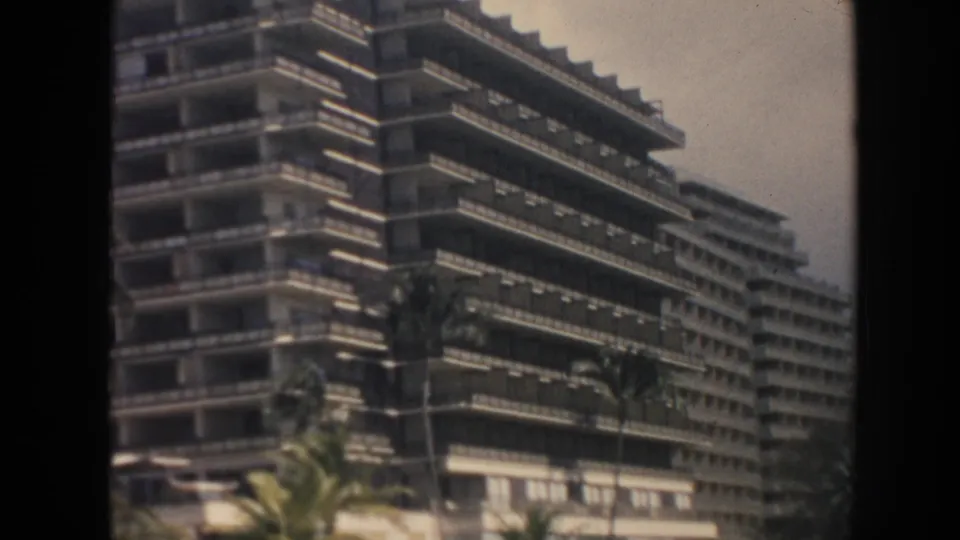 1961 Hawaii 8mm frame showing a large tiered hotel or apartment building with balconies, palms, and street-level frontage.